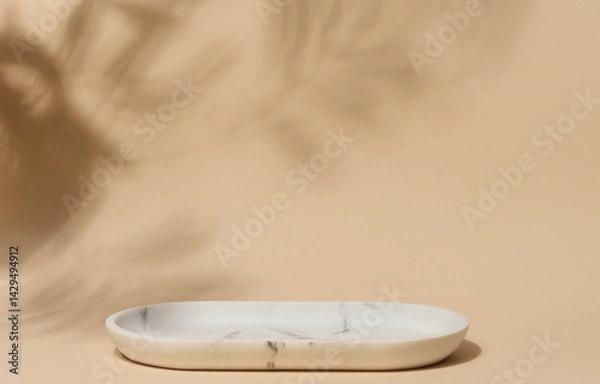 Fototapeta An empty, oval shaped soap dish made of white marble with gray veining sits on a beige background with soft, blurred leaf shadows