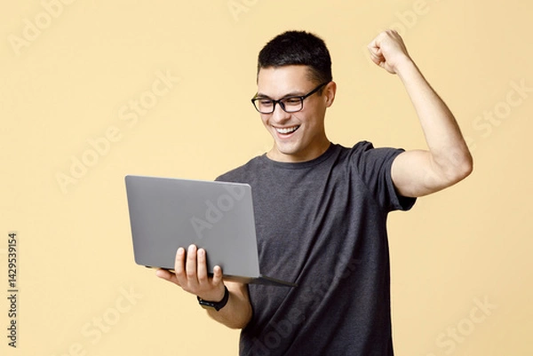 Fototapeta Great job, solving problem and win in game. Happy young guy student with glasses looks at laptop and expresses emotions of victory, isolated on beige background, studio shot