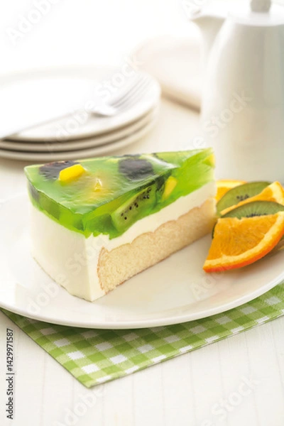 Fototapeta Close-up of a slice of kiwi cheesecake with a clear green jelly topping, kiwi slices, and orange wedges on a white plate.