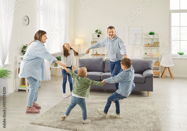 Fototapeta Happy large family holding hands and dancing in circle in spacious living room. Smiling parents and three children enjoying playful activity together, having fun and dancing in round dance at home.