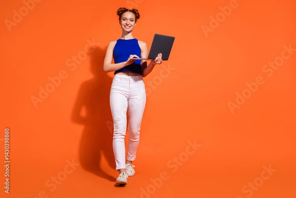 Fototapeta Youthful redheaded woman holding laptop, smiling on orange background, white pants and blue top, emphasizing casual fashion and technology