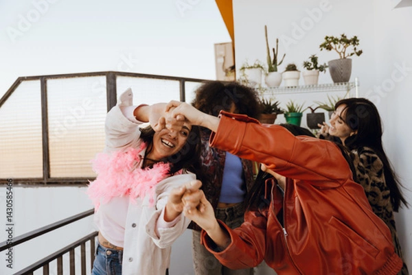 Fototapeta A group of joyful young women of different ethnicities in festive clothes dance and laugh together on a balcony at sunset, surrounded by potted plants. Concept of girls' party and friendship.