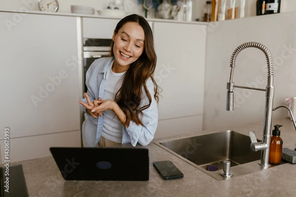 Obraz A calm European brunette woman aged 25, smiling widely while looking at her laptop as she stands next to the sink in a modern, minimalistic kitchen, talking to someone on videocall