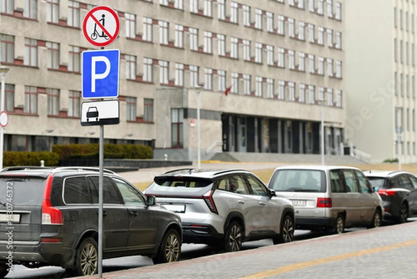 Fototapeta Row of parked vehicles along city sidewalk in front of government-style building with visible parking and restriction signs, no scooter zone warnings on cloudy day. Parking with multiple cars lined up