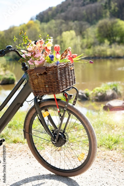 Fototapeta A bicycle with colorful flowers and baguette filled basket stands on a path in an idyllic spring landscape. Sunny bike tour backdrop for eco-friendly tourism, leisure, and lifestyle. Space for text.