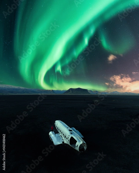 Obraz Abandoned DC-3 plane wreck on Solheimasandur beach in Iceland beneath vibrant green Northern Lights, with a person in red for scale and contrast.