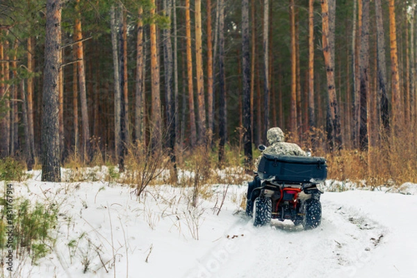 Fototapeta A man in camouflage is driving a four wheeler through a snowy forest