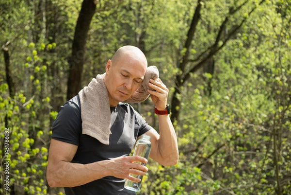 Obraz Sporty man wipes sweat from forehead, tired after running and jogging at forest path. Heat summer. Water balance.