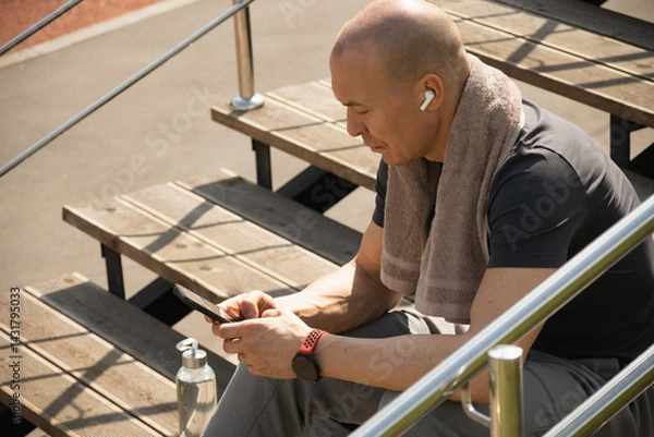 Obraz Athlete sitting on stairs and checking apps with health indicators, water balance after training. Relax after training. Man listens to music. Heat weather. Summer season. Close up.