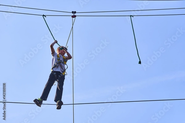 Fototapeta Cute school boy enjoying a sunny day in a climbing adventure activity park. Clear blue sky background. Children summer activities.
