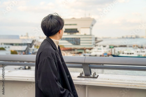 Fototapeta A Japanese man in his twenties wearing a black jacket walks along a seaside path in Minato Mirai, Yokohama, Kanagawa Prefecture, enjoying the breeze, city buildings, and the calm harbor atmosphere.