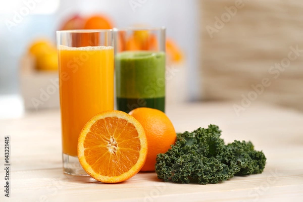 Obraz Orange juice and green vegetable juice in tall glasses with orange half kale and fruit basket in the background