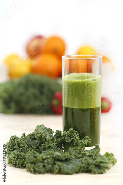 Obraz Green vegetable juice in a tall glass with a leaf of kale and a basket of fruits and vegetables in the background