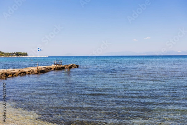 Obraz Tranquil seaside scene with clear blue water, a quaint rocky pier featuring a bench and flagpole, set against majestic mountains and vibrant coastal flora for relaxation.