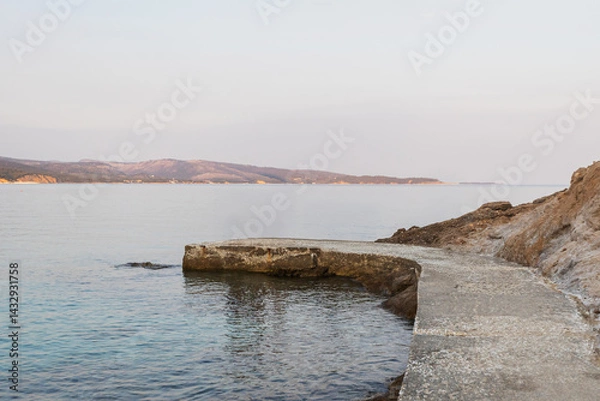 Obraz A winding concrete path follows a rugged rocky coastline and extends into the calm sea. Distant land is visible on the horizon under a clear sky, evoking solitude.
