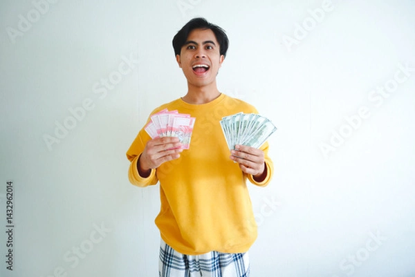Obraz Happy young Asian man holding Indonesian rupiah and US dollar bills with excited expression, perfect for finance and success themes
