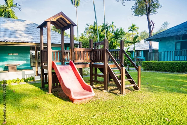 Fototapeta Wooden playground set with a red slide in a sunny backyard surrounded by tropical homes and lush greenery
