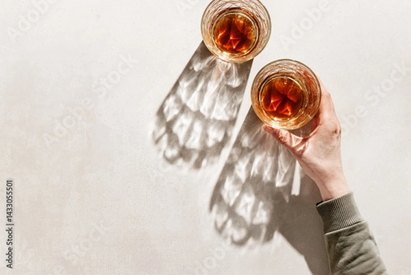 Obraz Two Whiskey Glasses with Shadows and sunlight Reflection on beige background, hand holding glass with alcohol, aesthetic top view summer still life, refraction at crystal shine cups, caustics