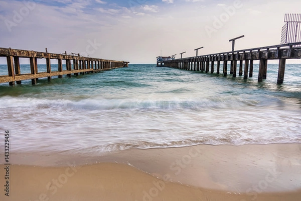 Fototapeta Rustic parallel piers stretch out into the ocean with gentle waves washing onto the sandy beach under a partly cloudy sky