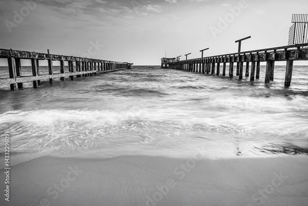 Fototapeta Black and white photo long exposure of rustic parallel piers stretch out into the ocean with gentle waves washing onto the sandy beach under a partly cloudy sky