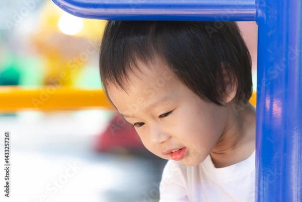 Obraz Asian young child gazes with curiosity, peering out from beneath a playful playground structure, a picture of innocent wonder and exploration. Closeup Curious kid boy.