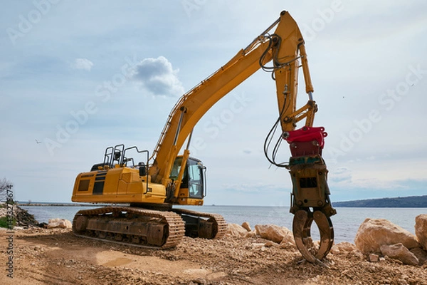 Fototapeta Yellow tracked excavator with hydraulic claw attachment working on a rocky coastal construction site near the sea under a partly cloudy sky