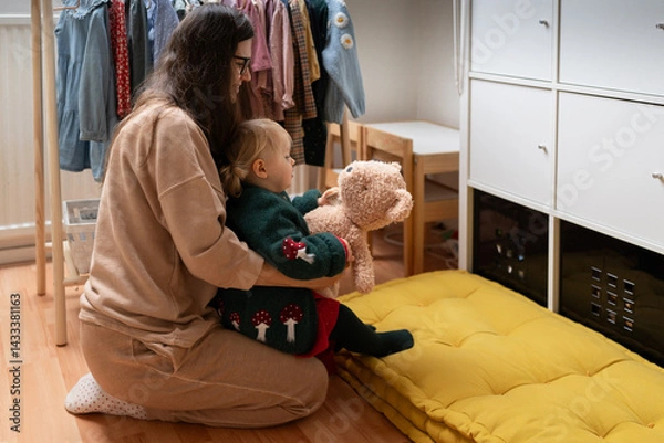 Obraz Authentic cozy moment of a mother and her little daughter hugging at home, surrounded by children's clothes and toys. Real life, real emotions, real love.