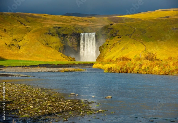 Obraz Skogafoss waterfall in Iceland cascades down a steep cliff, creating a powerful mist that forms a vibrant rainbow. The surrounding lush green landscape enhances its majestic beauty
