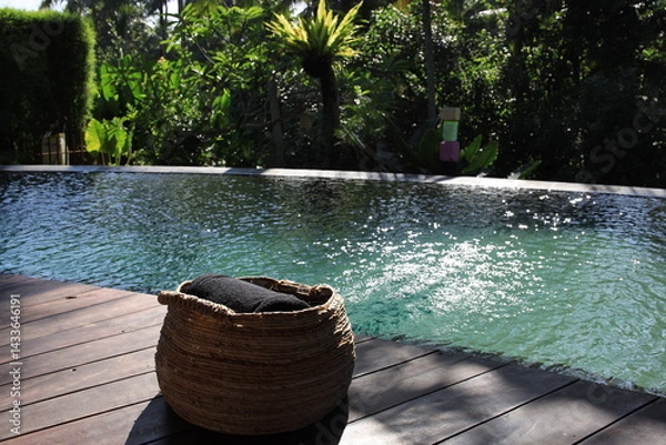 Fototapeta Basket with a black towel sits on a wooden deck next to a pool. The pool is surrounded by trees and has a calm, peaceful atmosphere