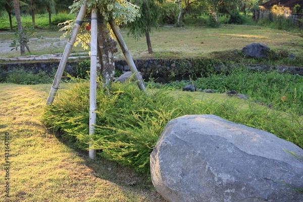 Fototapeta Large rock sits in a grassy field next to a tree. The rock is surrounded by a small bush and a metal pole. The scene is peaceful and serene, with the sun shining down on the grass and the rock