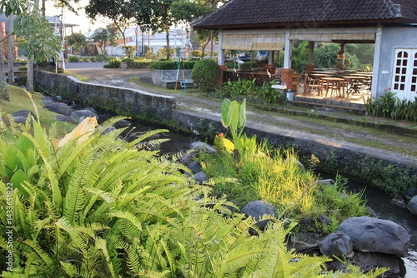 Fototapeta Lush green garden with a small stream running through it. The garden is surrounded by a stone wall and has a wooden pavilion in the background