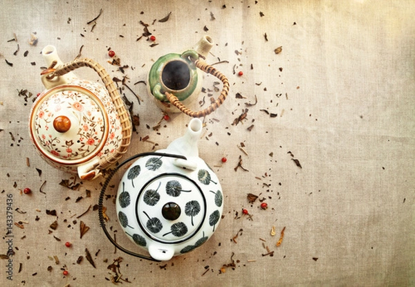 Fototapeta 3 various traditional ceramic teapots on a canvas background with copy space. Group of cute steaming clayware pots  with hot tea among tea leaves and berries scattered on fabric
