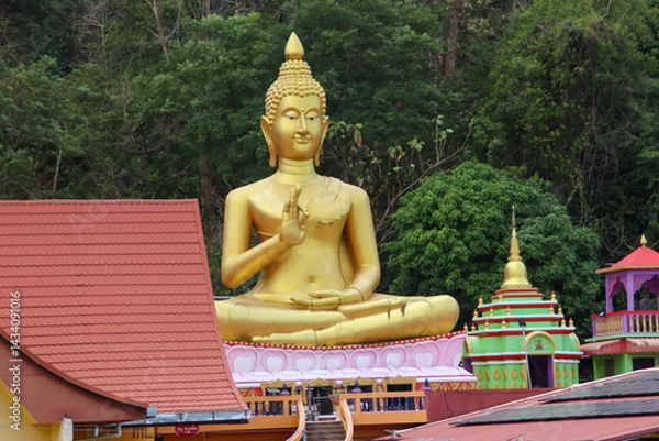 Fototapeta Golden Big Buddha statue stands near traditional Thai-style buildings, with lush green forest in the background. 