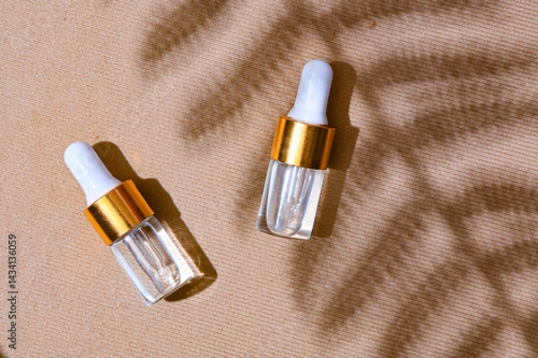 Fototapeta Two transparent dropper serum bottles with white caps and gold collars placed on a textured beige background with soft leaf shadows. Natural skincare branding concept with summer mood