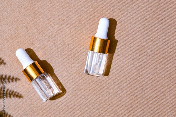 Fototapeta Two transparent dropper serum bottles with white caps and gold collars placed on a textured beige background with soft leaf shadows. Natural skincare branding concept with summer mood