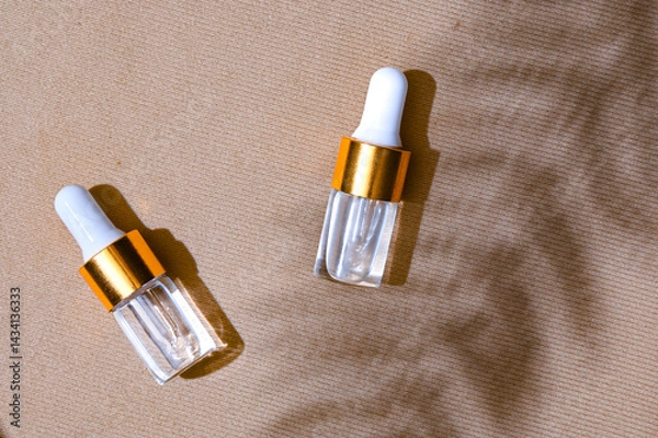 Fototapeta Two transparent dropper serum bottles with white caps and gold collars placed on a textured beige background with soft leaf shadows. Natural skincare branding concept with summer mood
