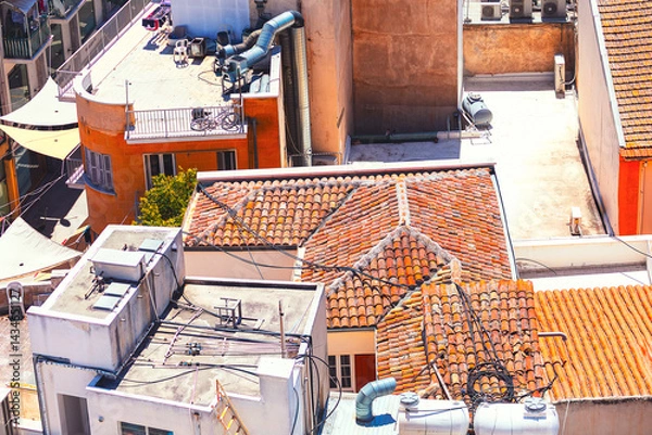 Obraz Aerial view shows a cluster of buildings with red-tiled roofs and modern rooftops, adorned with pipes, vents, and equipment. Scene includes a mix of orange and white structures under sunny sky