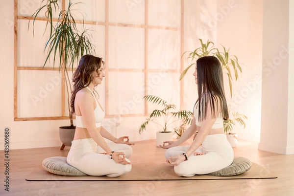 Fototapeta Two women sit cross-legged facing each other in soft light, connection through synchronized breath and mutual focus, connection deepens in tranquil yoga studio atmosphere bond captured composition 