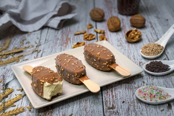 Fototapeta Bitten vanilla ice cream bar with chocolate and nut coating on ceramic plate. Rustic wooden background, surrounded by natural ingredients