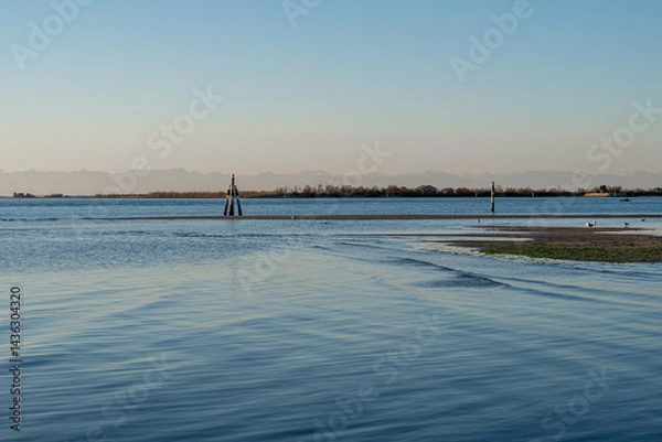 Fototapeta Lagoon Waters and Rocky Shoreline of Grado Framed by Snow-Capped Alpine Mountains and a Green Beacon under a Clear Blue Sky.