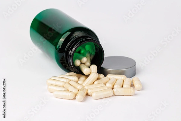 Obraz Spilled capsules from green bottle on white background, Close-up of beige capsules spilling out of a green plastic bottle onto a clean white background, representing supplements and healthcare.  
