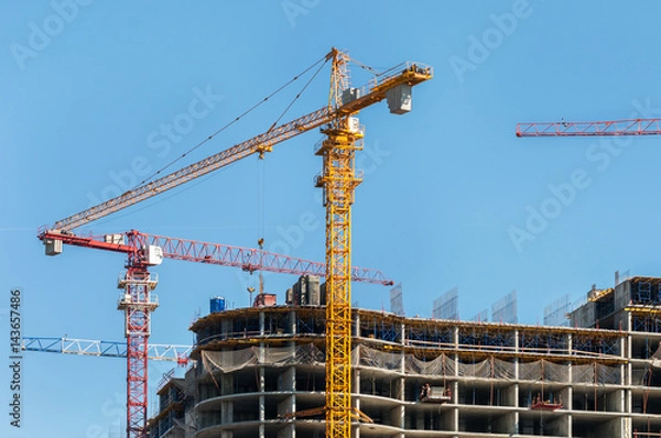 Fototapeta Construction site. Construction cranes and high-rise building under construction against blue sky.