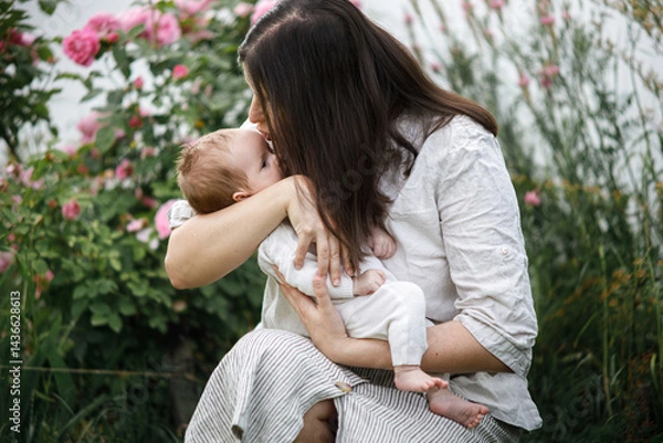 Fototapeta Mother kissing and hugging cute newborn baby among flowers in the garden. Love, care and family in countryside concept. Happy Mothers day! Adoption and maternity
