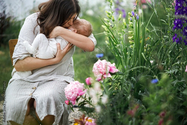 Fototapeta Mother kissing and hugging cute newborn baby among flowers in the garden. Love, care and family in countryside concept. Happy Mothers day! Adoption and maternity