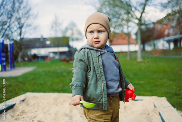 Fototapeta A boy of 2 years old in a spring jacket and hat plays with sand in a sandbox. A boy plays on a playground with a shovel in the sand. A child's walk in the fresh air