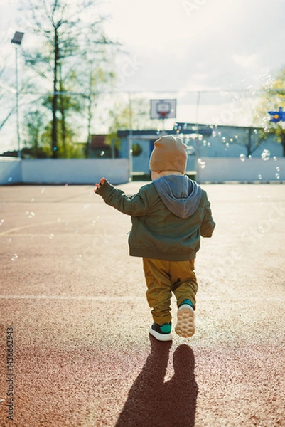 Fototapeta Cheerful 2 year old boy playing outside in summer in a green jacket with soap bubbles. Toddler boy running outside and playing with bubbles.