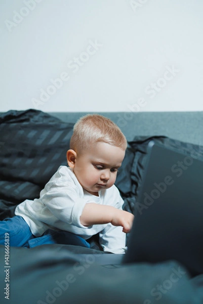 Fototapeta A 2-year-old boy sits in bed and presses buttons on a laptop. The child plays with a laptop.