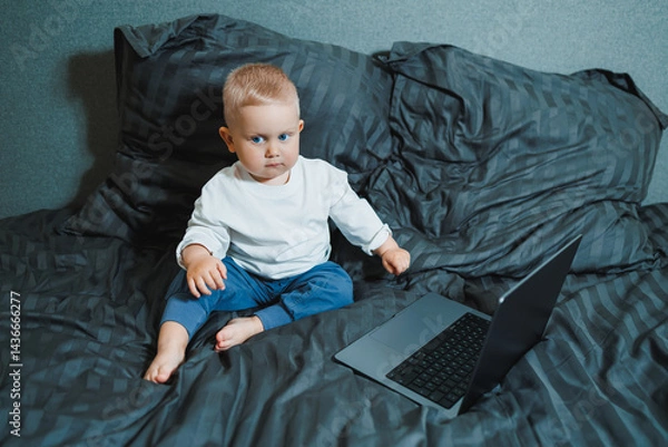 Fototapeta A 2-year-old boy sits in bed and presses buttons on a laptop. The child plays with a laptop.