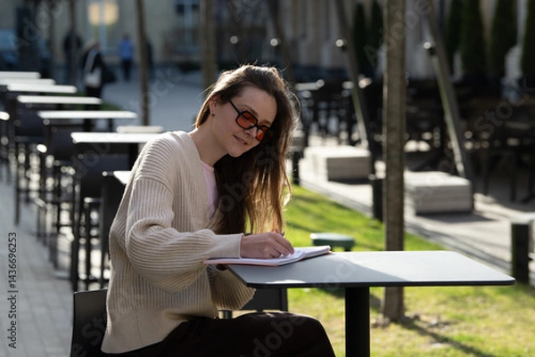 Fototapeta A young woman sits at a black table outdoors, writing in her notebook under soft afternoon sunlight