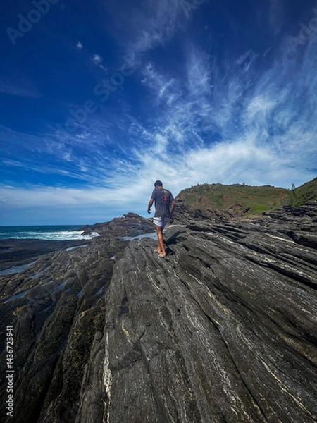 Obraz A young man walks on the rocks along the shore of a beach in the town of Búzios, the main tourist destination in the lakes region of Rio de Janeiro.